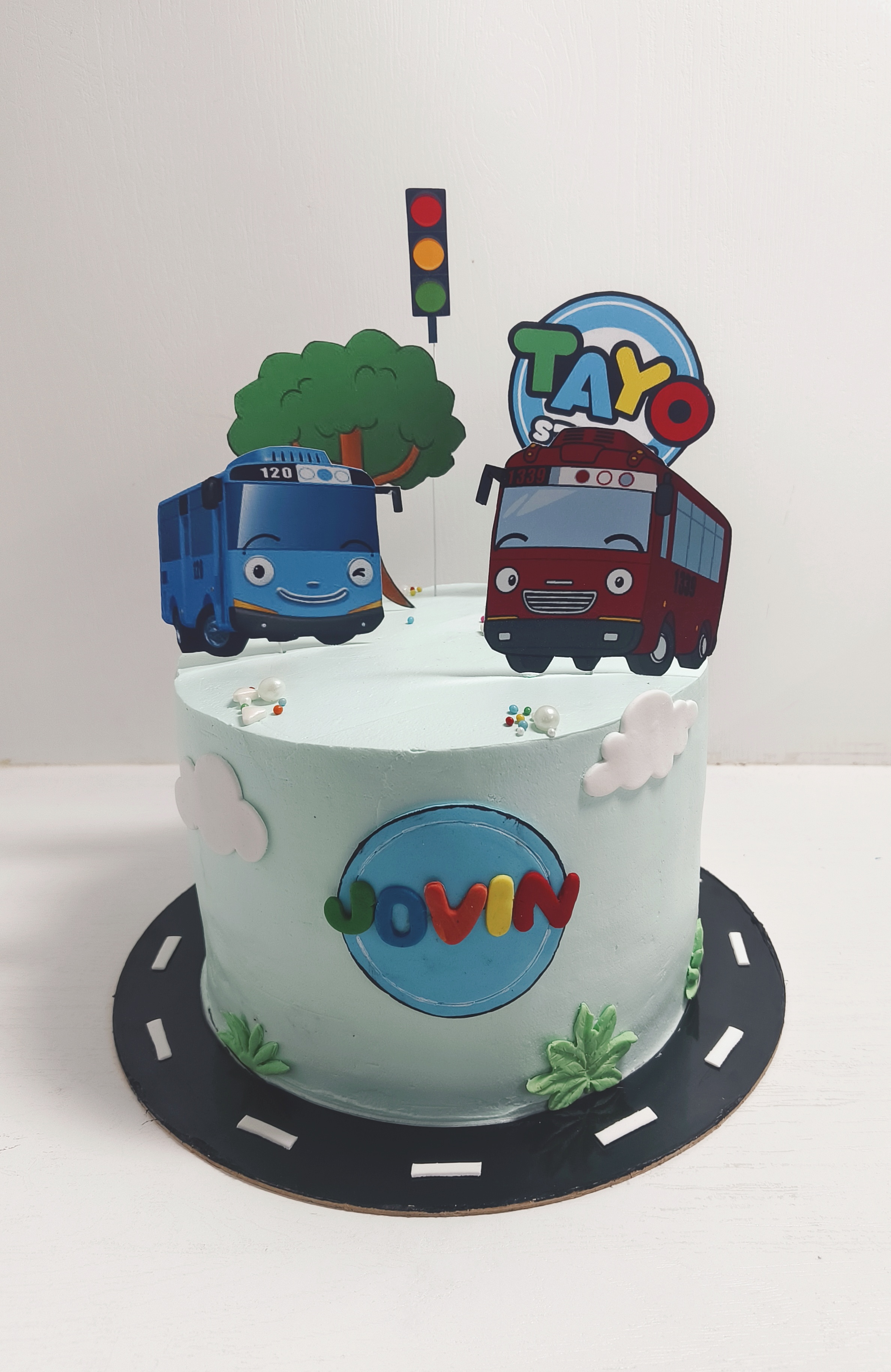 Birthday cake with cartoon bus toppers, clouds, and a road around the base